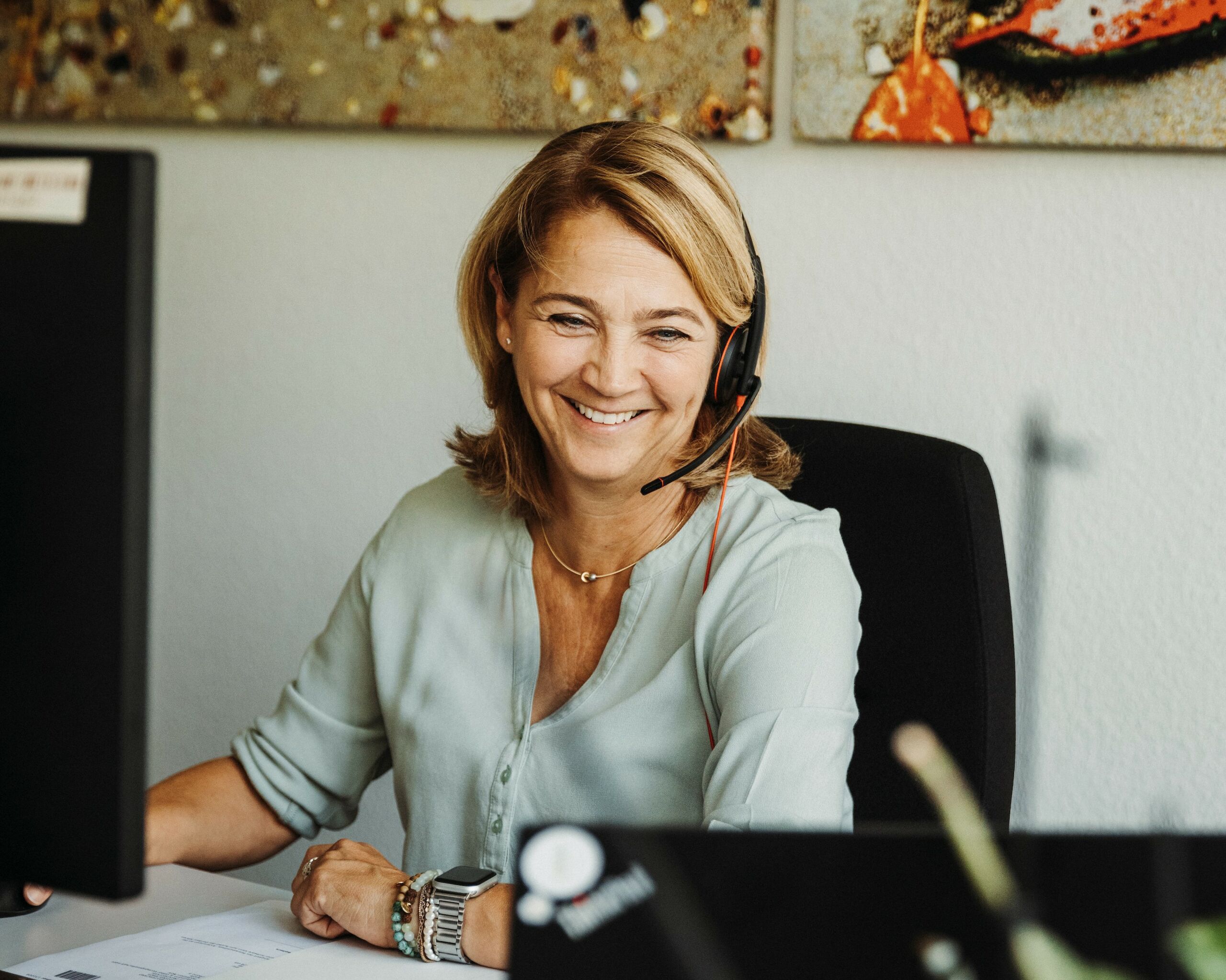 Eine lachende Mitarbeiterin sitzt an ihrem Schreibtisch mit einem Headset